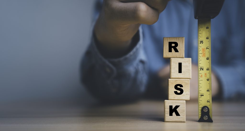 Close-up concept image of a person measuring a stack of wooden blocks spelling “RISK” with a yellow tape measure, representing the need to identify, assess and control dangerous substances and explosive atmosphere risks during a DSEAR risk assessment.