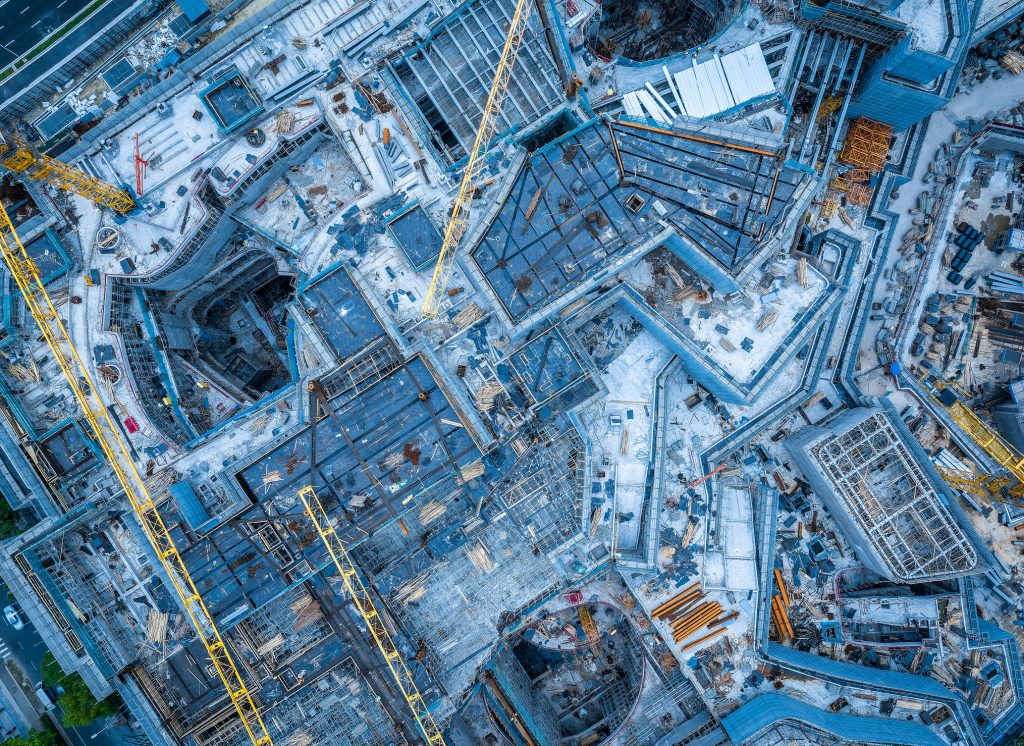 Aerial view of a large, active construction site containing partially completed buildings, scaffolding, materials and several tower cranes, illustrating the range of machinery and site activities that can contribute to workplace and environmental noise during summer construction projects.